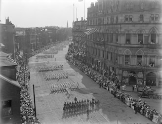 A large parade with uniformed participants marching in formation down a wide cobblestone street. Crowds of onlookers line the sidewalks on both sides, and flags and banners decorate the buildings. The architecture features large multi-story buildings with arched windows. Automobiles are parked along the side.