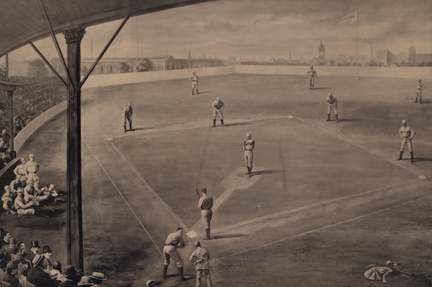 A vintage sepia-toned illustration of a baseball game in progress, showing players on the field in various positions. The scene includes a crowd of spectators in the stands, dressed in early 20th-century attire. A flag with a city's name is visible in the background, along with an urban skyline. The perspective provides a comprehensive view of the baseball diamond and the surrounding stadium.