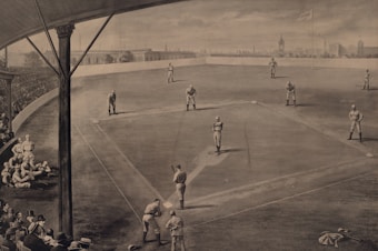 A vintage sepia-toned illustration of a baseball game in progress, showing players on the field in various positions. The scene includes a crowd of spectators in the stands, dressed in early 20th-century attire. A flag with a city's name is visible in the background, along with an urban skyline. The perspective provides a comprehensive view of the baseball diamond and the surrounding stadium.
