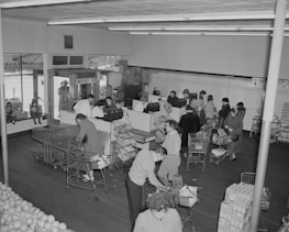 A busy grocery store with customers standing in line at the checkout counters. Shoppers with carts are filled with various grocery items. The store has a section with stacked goods and several people are being attended by clerks behind the counters. Natural light enters through the front glass windows.