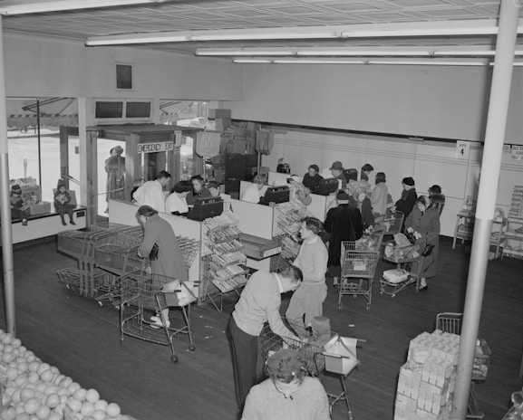 A busy grocery store with customers standing in line at the checkout counters. Shoppers with carts are filled with various grocery items. The store has a section with stacked goods and several people are being attended by clerks behind the counters. Natural light enters through the front glass windows.