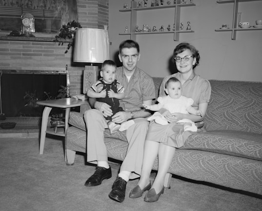 A family of four is seated on a couch in a living room. The father holds a young boy in his lap, while the mother holds an infant girl. The room features a fireplace with a decorative clock on the mantel, a mid-century style lamp, a side table with a radio, and shelves with various figurines and decorations. The family appears happy and well-dressed, with the father and son in button-up shirts and slacks, and the mother wearing glasses, a blouse, and a skirt.