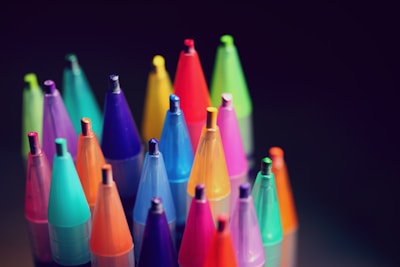 An arrangement of colorful crayons stands vertically against a dark background. Each crayon is encased in a translucent plastic container, with vibrant colors including shades of blue, green, red, purple, yellow, and orange. The lighting highlights the tip of each crayon, creating a visually appealing contrast.