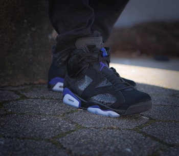 A shoe featuring a stylish and modern design is placed on a textured stone pathway, showcasing a blend of black, purple, and white colors. The shoe's material includes a mix of textures and patterns, and it is worn by someone wearing dark pants.