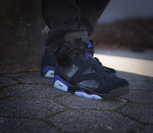 A shoe featuring a stylish and modern design is placed on a textured stone pathway, showcasing a blend of black, purple, and white colors. The shoe's material includes a mix of textures and patterns, and it is worn by someone wearing dark pants.