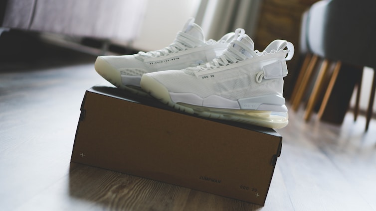 A pair of white high-top sneakers is placed on top of a brown shoebox. The sneakers have intricate detailing and visible air cushions in the sole. The scene is set in a bright room with a wooden floor, partially visible furniture, and soft lighting.