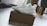 A pair of white high-top sneakers is placed on top of a brown shoebox. The sneakers have intricate detailing and visible air cushions in the sole. The scene is set in a bright room with a wooden floor, partially visible furniture, and soft lighting.