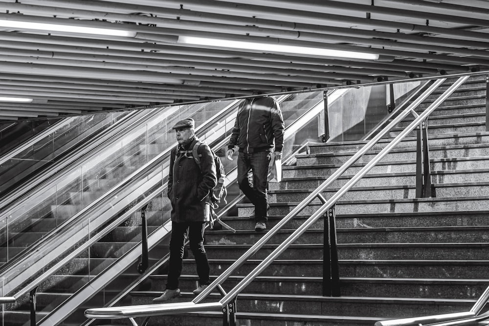 grayscale photo of man in jacket and pants walking down the stairs