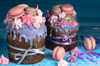 Two elaborately decorated cakes with purple icing and pastel-colored meringues and macarons. Both cakes are wrapped with festive ribbons. The scene is vibrant with a scattering of meringues and macarons on the blue surface.