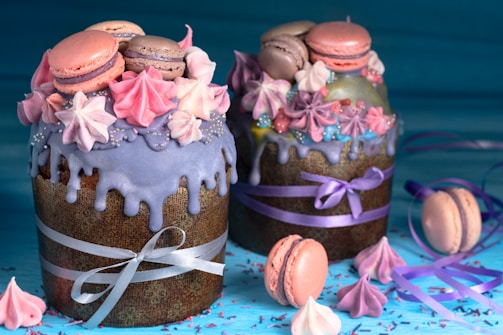 Two elaborately decorated cakes with purple icing and pastel-colored meringues and macarons. Both cakes are wrapped with festive ribbons. The scene is vibrant with a scattering of meringues and macarons on the blue surface.