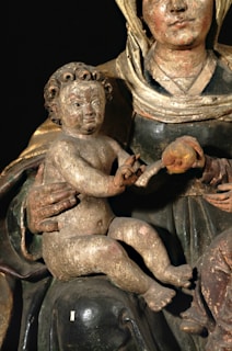 A vintage wooden sculpture depicting a seated woman holding an infant with curly hair. The woman is dressed in traditional attire with a headscarf and a dark-coloured robe. The infant is unclothed with visible signs of age and wear on the wood. The figures have a serene expression.