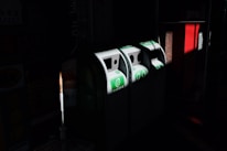 Three recycling bins are positioned side by side in a dimly lit area, with green signage indicating their purpose. Light highlights the bins, casting shadows around them. Nearby, a red structure is visible, contrasting with the dark surroundings.