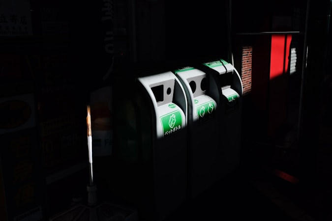 Three recycling bins are positioned side by side in a dimly lit area, with green signage indicating their purpose. Light highlights the bins, casting shadows around them. Nearby, a red structure is visible, contrasting with the dark surroundings.