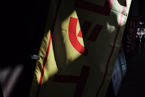 A sign or banner with red lettering set against a yellow background is partially illuminated by sunlight. The surrounding area is in shadow, giving contrast to the illuminated section of the banner. In the background, there are other signs and a dimly lit street or hallway.