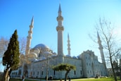 A large mosque with multiple tall minarets and a prominent central dome. The building is surrounded by leafless trees and a few evergreen trees in a well-maintained garden setting. Several people are walking and gathering near the structure under a clear blue sky.