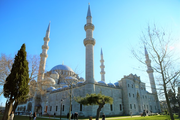 A large mosque with multiple tall minarets and a prominent central dome. The building is surrounded by leafless trees and a few evergreen trees in a well-maintained garden setting. Several people are walking and gathering near the structure under a clear blue sky.