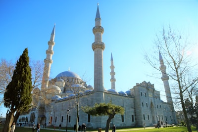 A large mosque with multiple tall minarets and a prominent central dome. The building is surrounded by leafless trees and a few evergreen trees in a well-maintained garden setting. Several people are walking and gathering near the structure under a clear blue sky.