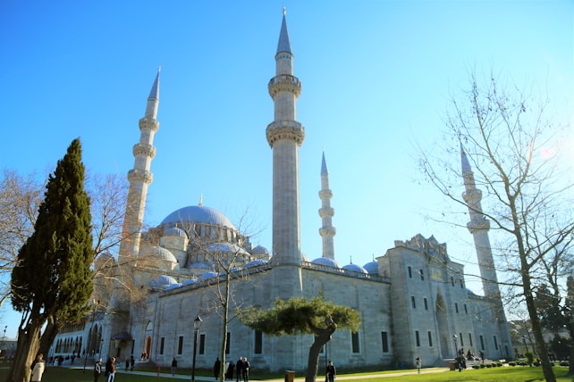 A large mosque with multiple tall minarets and a prominent central dome. The building is surrounded by leafless trees and a few evergreen trees in a well-maintained garden setting. Several people are walking and gathering near the structure under a clear blue sky.