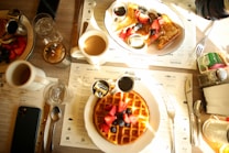 A table set for breakfast with plates featuring a waffle topped with fresh berries and another dish with french toast, strawberries, and blueberries. Accompanied by mugs of coffee and cups, silverware, and small metal syrup containers. The table is organized with menus beneath the plates and various condiments.