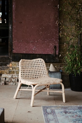 A woven rattan chair is positioned on a concrete or stone floor. It's set against a rustic backdrop with aged wooden shutters and a wall made of bricks or stone. To the right, there is a tall plant in a black pot, and a small rug is partially visible in the foreground.