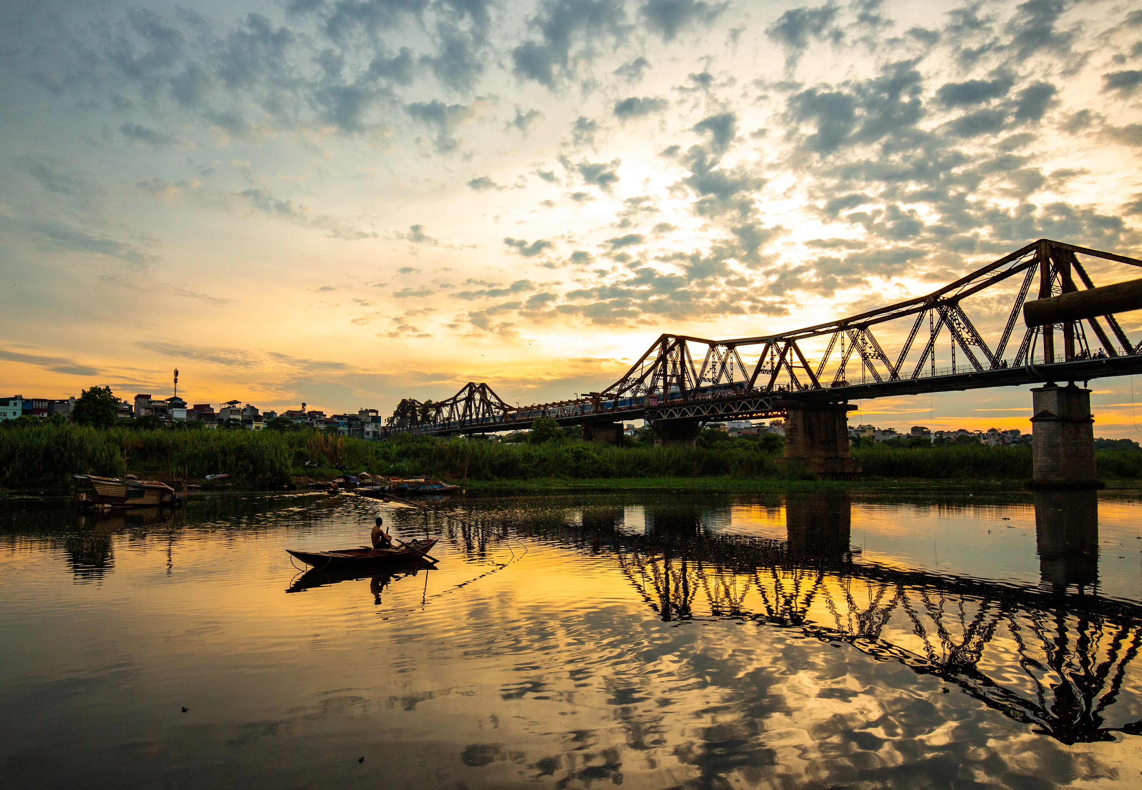 Long Bien Bridge silhouetted at golden sunset with a boat on the reflecting river below.