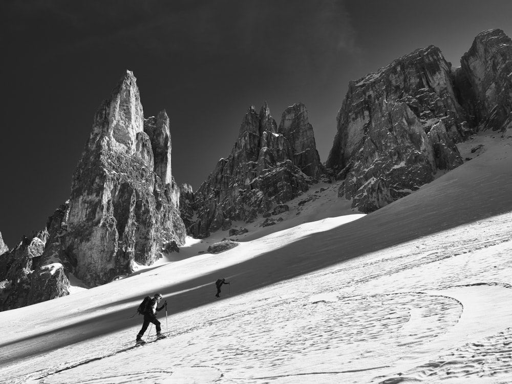grayscale photo of man walking on snow covered field near rocky mountain