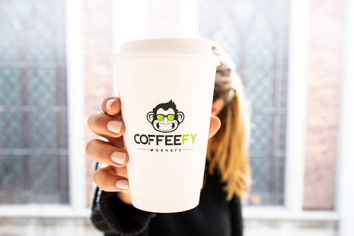 A close-up of a white paper coffee cup with a distinctive logo featuring a cartoon monkey wearing sunglasses. The word 'COFFEEFY' is printed on the cup, held by a person with neatly manicured nails. The background is blurred, suggesting a setting with large windows and a bright ambiance.