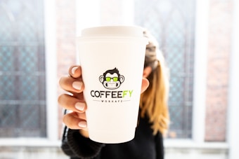A close-up of a white paper coffee cup with a distinctive logo featuring a cartoon monkey wearing sunglasses. The word 'COFFEEFY' is printed on the cup, held by a person with neatly manicured nails. The background is blurred, suggesting a setting with large windows and a bright ambiance.