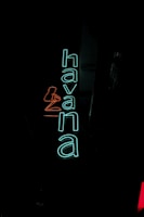 Bright neon sign with the word 'havana' in turquoise and an outline of a seated woman in red. The sign is against a dark background, making the colors pop vividly.