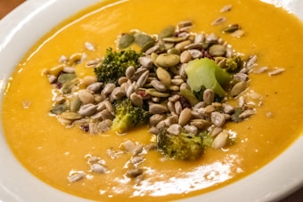 A bowl of creamy orange soup, garnished with a variety of seeds and small pieces of broccoli. The soup appears smooth and rich in texture, with the seeds and broccoli providing a contrasting texture.