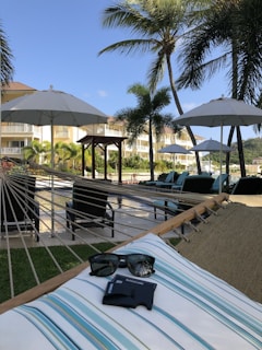 A hammock stretches across a grassy area under the shade of tall palm trees, with a view of poolside lounge chairs and umbrellas. In the foreground, a pair of sunglasses and a cardholder rest on a striped cushion. Tropical plants and a multi-story building are visible in the background under a clear blue sky.