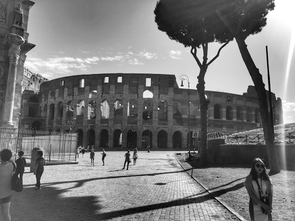 The black and white image captures the iconic ancient Roman Colosseum, with its archways and historical architecture illuminated by sunlight. Visitors are scattered around the cobblestone plaza, casting long shadows. A large tree and lampposts add depth to the scene.