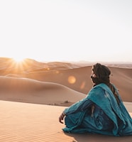 woman in blue hoodie sitting on sand during sunset