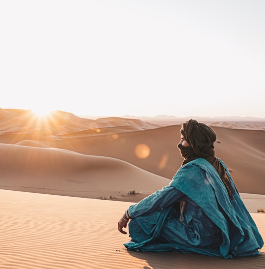 woman in blue hoodie sitting on sand during sunset in Merzouga Morocco