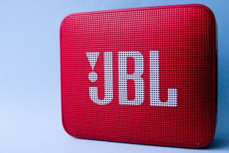 A red portable Bluetooth speaker with a perforated front and large logo in white lettering. The surface is textured with small, evenly spaced holes. It is set against a plain light blue background.