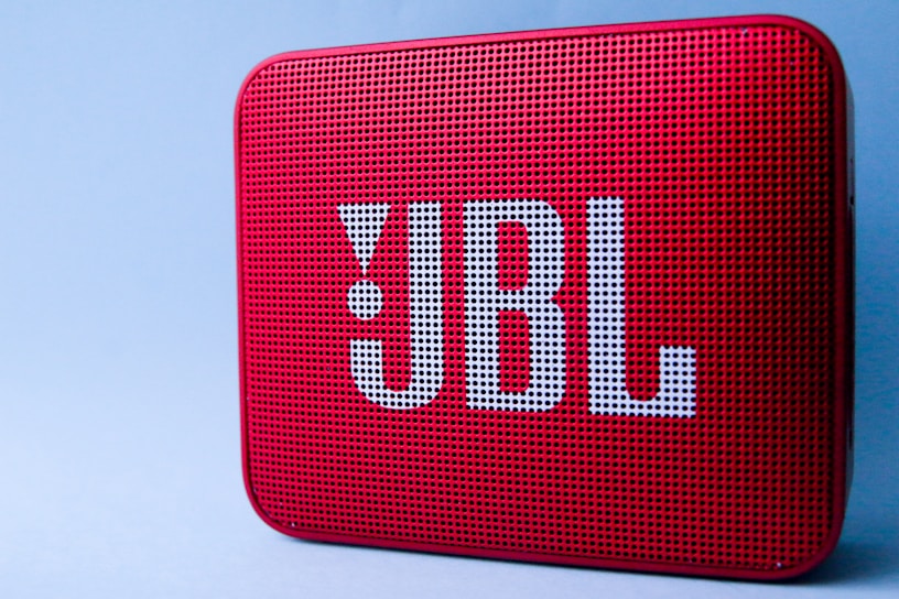 A red portable Bluetooth speaker with a perforated front and large logo in white lettering. The surface is textured with small, evenly spaced holes. It is set against a plain light blue background.