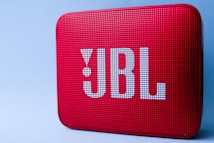 A red portable Bluetooth speaker with a perforated front and large logo in white lettering. The surface is textured with small, evenly spaced holes. It is set against a plain light blue background.