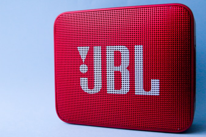 A red portable Bluetooth speaker with a perforated front and large logo in white lettering. The surface is textured with small, evenly spaced holes. It is set against a plain light blue background.