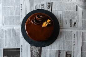 A round chocolate cake with a smooth, glossy finish is placed on a dark slate platter. Decorative chocolate swirls and yellow edible flowers are perched on top. The background consists of newspaper pages laid out flat.