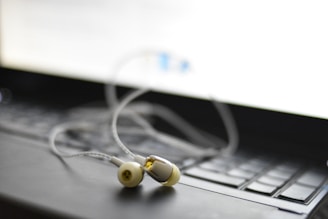 A pair of earphones rests on a laptop keyboard. The focus is on the earphones, with their white and yellow details visible. The laptop's screen emits a bright light, creating a soft blur in the background.