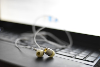 A pair of earphones rests on a laptop keyboard. The focus is on the earphones, with their white and yellow details visible. The laptop's screen emits a bright light, creating a soft blur in the background.