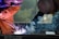 Two workers are engaged in welding, creating bright sparks and smoke. They are wearing protective clothing and helmets, amidst an industrial setting. The environment appears focused and intense, with visible safety gear.
