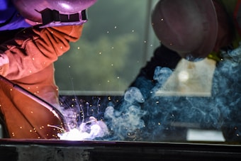 Two workers are engaged in welding, creating bright sparks and smoke. They are wearing protective clothing and helmets, amidst an industrial setting. The environment appears focused and intense, with visible safety gear.