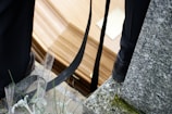 A close-up view of a coffin resting on a stone or concrete surface, with black straps and parts of two people wearing dark-colored shoes nearby. The area is sprinkled with water droplets and there are some green plants and a granite surface visible.