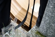 A close-up view of a coffin resting on a stone or concrete surface, with black straps and parts of two people wearing dark-colored shoes nearby. The area is sprinkled with water droplets and there are some green plants and a granite surface visible.