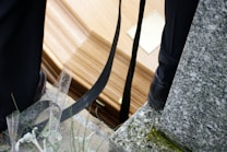 A close-up view of a coffin resting on a stone or concrete surface, with black straps and parts of two people wearing dark-colored shoes nearby. The area is sprinkled with water droplets and there are some green plants and a granite surface visible.