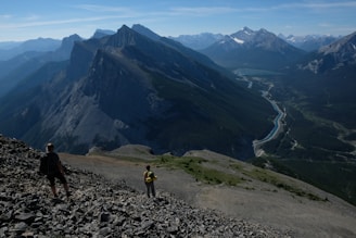 Two hikers traverse a rocky mountainside, offering a sweeping view of jagged peaks and a winding river below. The horizon reveals an expansive range of mountains under a clear blue sky.