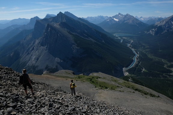 Two hikers traverse a rocky mountainside, offering a sweeping view of jagged peaks and a winding river below. The horizon reveals an expansive range of mountains under a clear blue sky.