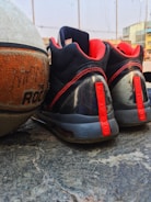 A pair of black and red athletic shoes is placed next to a worn-out basketball on a concrete surface. The laces and accents on the shoes are red, while the basketball has an orange texture with visible branding. The background includes a blurred part of a building and a clear sky, indicating an outdoor setting.