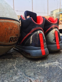 A pair of black and red athletic shoes is placed next to a worn-out basketball on a concrete surface. The laces and accents on the shoes are red, while the basketball has an orange texture with visible branding. The background includes a blurred part of a building and a clear sky, indicating an outdoor setting.
